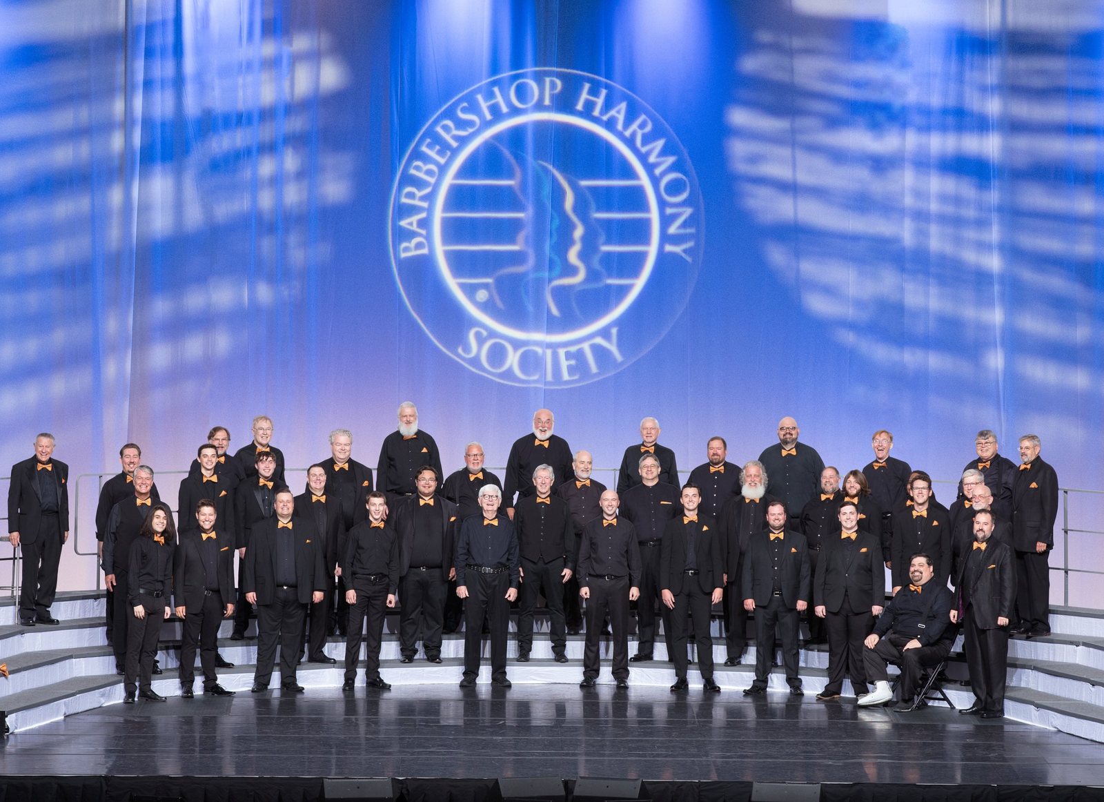 The chorus in black suits and orange bow ties standing on the Barbershop Harmony Society International stage in Charlotte, North Carolina, 2022.