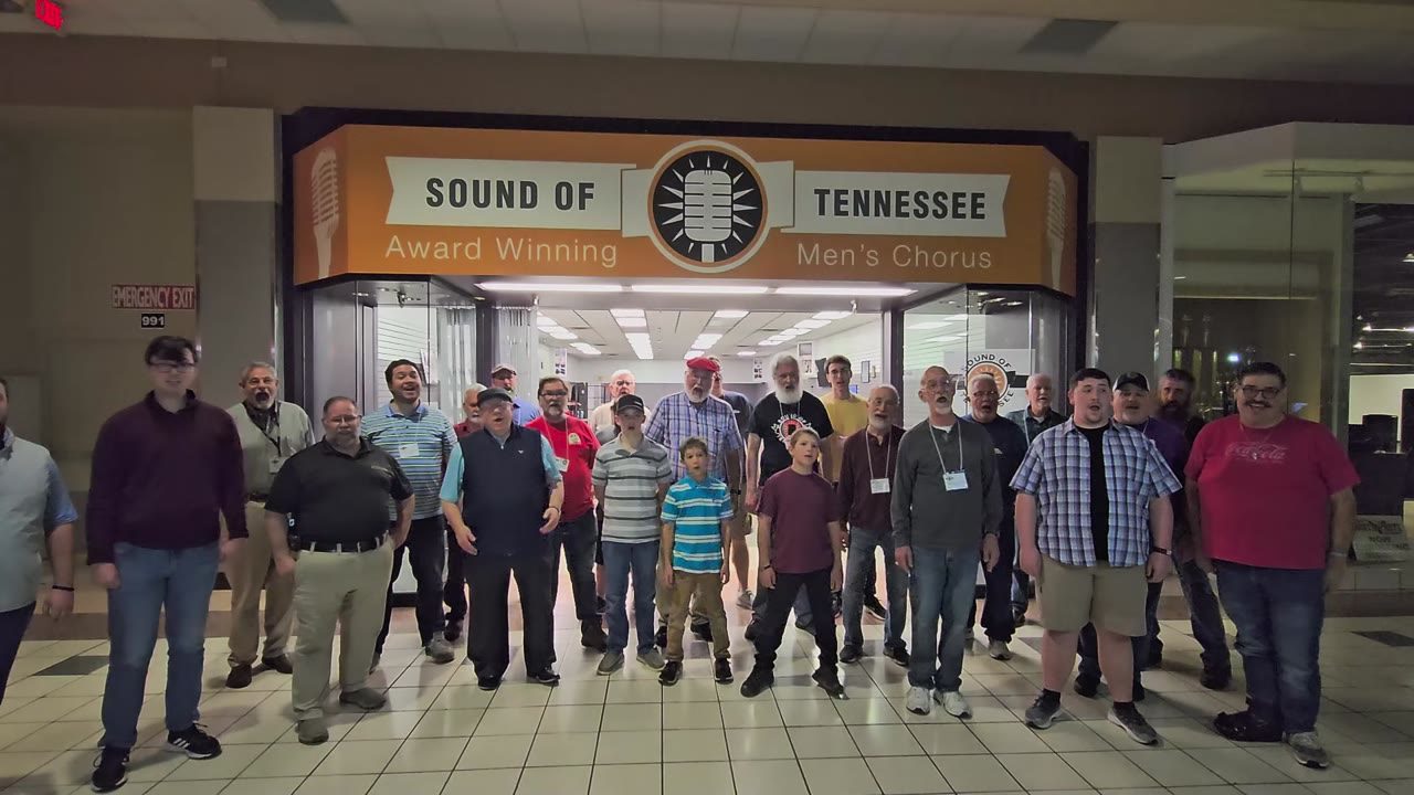 About forty men and a few boys, singing together in a mall corridor in front of a big orange sign that reads Sound of Tennessee, Award Winning Men's Chorus.